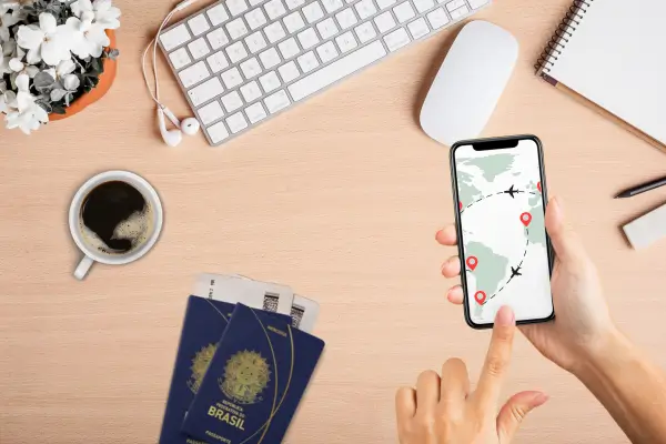 Pessoa organizando viagem internacional usando celular, passaportes brasileiros e itens de planejamento sobre mesa clara